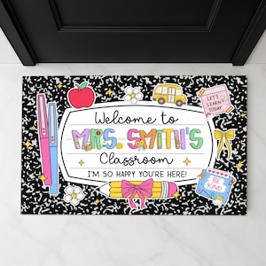 May include: A classroom-themed doormat with a black and white notebook paper design. The mat features colorful illustrations of school supplies, including pens, an apple, a school bus, and a pencil with a pink bow. Text reads "Welcome to Mrs. Smith's Classroom."