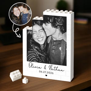May include: A white building block display stand featuring a black and white photograph of a couple. The text Olivia & Nathan and the date 06.27.2026 are printed below the image, followed by a small heart icon. Two loose blocks sit nearby.