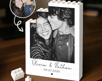 Può includere: Un espositore a blocchi di costruzione bianchi con una fotografia in bianco e nero di una coppia. Il testo Olivia & Nathan e la data 27.06.2026 sono stampati sotto l'immagine, seguiti da un piccolo cuore. Due blocchi sciolti sono vicini.