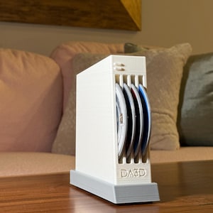 May include: A white 3D-printed CD storage case holding several CDs. The case has a rectangular shape with vertical slots and a gray base. The text "DA3D" is printed on the front. The CDs have blue, red, and silver labels.