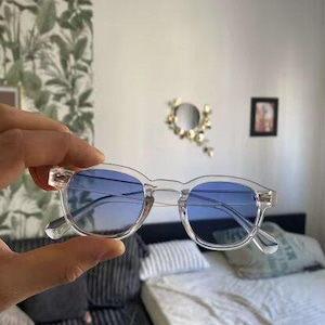 Peut inclure: Lunettes de soleil à monture transparente avec des verres bleus. Les lunettes sont tenues dans une main.