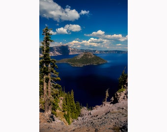 May include: A panoramic view of Crater Lake in Oregon, with a small island in the center of the lake. The lake is a deep blue color, and the sky is a bright blue with white clouds. The view is from a high vantage point, with trees in the foreground.