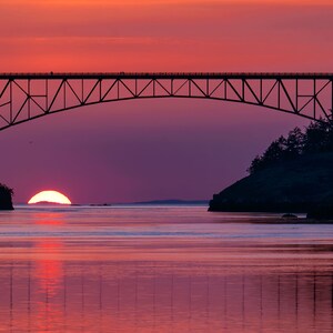 May include: A silhouette of a bridge over a body of water with a setting sun in the background. The sky is a vibrant pink and purple.