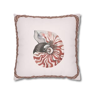 Puede incluir: Una almohada blanca con un borde de cuerda marrón presenta una ilustración de acuarela de una concha de nautilus con rayas rojas y blancas.