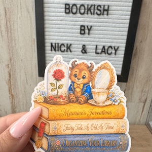 May include: A die-cut sticker featuring a stack of three books titled Maurice's Inventions, Fairy Tale As Old As Time, and Organizing Your Library. Atop the books sits a small creature in a blue coat, a red rose in a glass dome, and a teacup.