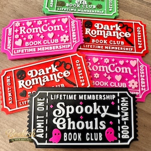 May include: A collection of rectangular ticket-shaped plaques featuring raised text and icons. The plaques include labels for RomCom, Dark Romance, and Spooky Ghouls book clubs, with details like Admit One, Lifetime Membership, and unique serial numbers.