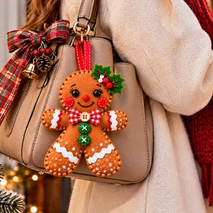 May include: A beige leather handbag adorned with a festive gingerbread man ornament featuring a red and white checkered bowtie, green buttons, and holly leaves. A plaid ribbon bow with a small gold bell is attached to the bag handle.