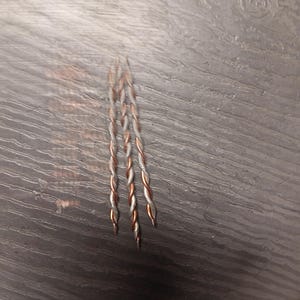 May include: Three twisted copper and silver wires are arranged vertically against a dark gray wood-grain surface. The wires are tightly braided, with the copper sections appearing reddish-brown and the silver sections a bright metallic color. The ends of the wires are pointed.