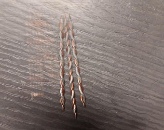 May include: Three twisted copper and silver wires are arranged vertically against a dark gray wood-grain surface. The wires are tightly braided, with the copper sections appearing reddish-brown and the silver sections a bright metallic color. The ends of the wires are pointed.