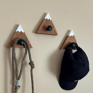 May include: Three wooden mountain-shaped hooks with black knobs. The hooks are painted white at the top to resemble snow-capped mountains. A rope is hanging from one hook and a black baseball cap is hanging from another.