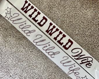 May include: Two white sashes with the words "Wild Wife" printed in a dark red, western-style font. One sash has the words "Wild Wild" above "Wife". Both sashes have star graphics.