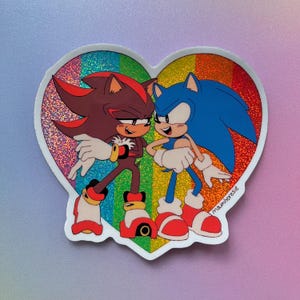 May include: A heart-shaped sticker with a rainbow background features Sonic the Hedgehog and Shadow the Hedgehog. Sonic is blue with a red and white shoe, and Shadow is black with a red and yellow shoe. The sticker has the text "mamiheresie" in the bottom right corner.