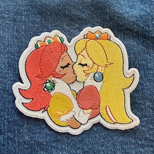 Könnte beinhalten: Gestickter Aufnäher mit zwei Comic-Prinzessinnen mit roten und gelben Haaren, die Kronen und Kleider tragen und sich küssen. Die Prinzessinnen haben braune Augen und sind auf einem weißen Hintergrund dargestellt.