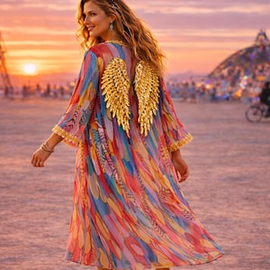 Puede incluir: Un vestido colorido y fluido con alas de ángel doradas en la espalda. El vestido presenta una vibrante mezcla de colores arcoíris. El fondo muestra un paisaje desértico con estructuras y personas, bajo un cielo de atardecer.