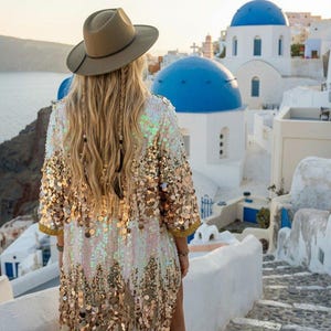 May include: A woman wearing a sequined, gold and white kimono-style jacket and a brown hat, standing on a stone pathway. The background features white buildings with blue domes, typical of Santorini, Greece.