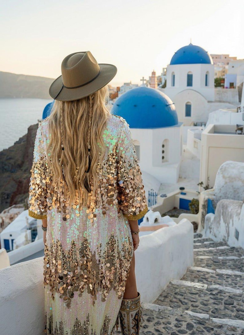May include: A woman wearing a sequined, gold and white kimono-style jacket and a brown hat, standing on a stone pathway. The background features white buildings with blue domes, typical of Santorini, Greece.