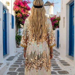 May include: A person wearing a long, sequined robe with gold and iridescent sequins, standing in a cobblestone alley. The robe has a gold trim and is paired with a black headband. The background includes white buildings, blue doors, and pink flowers.