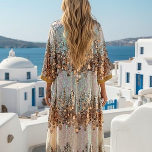 May include: A long, shimmering sequin dress with a gradient of colours from silver to gold. The dress has three-quarter sleeves with gold trim and is worn outdoors with a scenic background of white buildings and blue water.