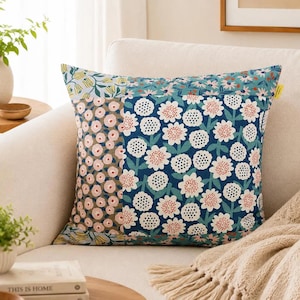 K&ouml;nnte beinhalten: Ein quadratisches Zierkissen mit Patchwork-Design und verschiedenen Blumenmustern. Der Stoff zeigt eine Mischung aus Dunkelblau, Hellrosa und Gelb mit stilisierten Blumenmotiven, darunter wei&szlig;e Bl&uuml;tenbl&auml;tter mit gepunkteten Mitten.