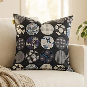 May include: A square throw pillow featuring a black background with a repeating pattern of circles. Each circle contains different designs, including floral motifs, plaid, and striped patterns in shades of blue, gray, white, and black.