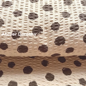 May include: Close-up of a textured fabric with a waffle weave pattern. The fabric is beige with large, dark brown polka dots. The text "Atelier Dorothy" is printed in white on the fabric.