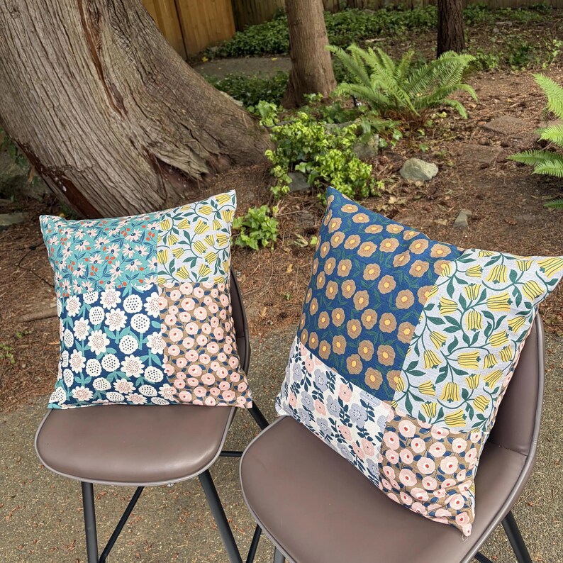 K&ouml;nnte beinhalten: Zwei quadratische Dekokissen mit einem Patchwork-Design aus Blumenmustern. Der Stoff zeigt verschiedene botanische Drucke in Blau-, Gelb-, Rosa- und Wei&szlig;t&ouml;nen mit sich wiederholenden Motiven von kleinen Blumen, Tulpen und kreisf&ouml;rmigen Bl&uuml;ten.