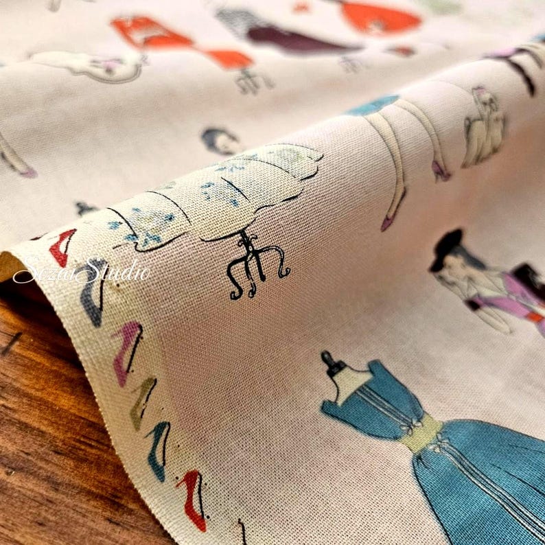 K&ouml;nnte beinhalten: Ein hellbeiger Stoff mit einem sich wiederholenden Muster aus verschiedenen bunten Kleidern, Schneiderpuppen und hochhackigen Schuhen. Die Illustrationen sind in Blau-, Rot-, Rosa- und Schwarzt&ouml;nen auf einer strukturierten Webstoffoberfl&auml;che.