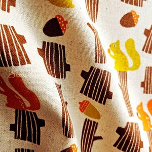 Pu&ograve; includere: Tessuto color crema con un motivo ripetuto di tronchi d'albero marroni, ghiande e scoiattoli gialli. Il design ha un'atmosfera vintage e autunnale, con un aspetto leggermente strutturato.