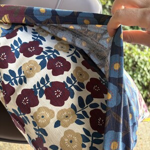 May include: A fabric pillow cover featuring a floral pattern with maroon, mustard yellow, and dark blue flowers on a cream background. The edge of the fabric is folded back to reveal a contrasting blue floral print lining with yellow centers.