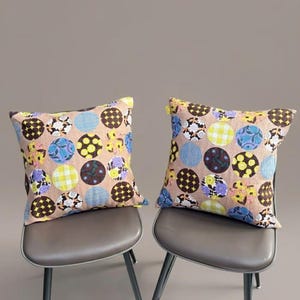 K&ouml;nnte beinhalten: Zwei quadratische Kissen mit einem beigen Grundstoff, der mit einem sich wiederholenden Muster aus bunten Kreisen verziert ist. Die Kreise enthalten verschiedene Designs, darunter gelbe Blumen, blaue Streifen, braune Gitter und violette Blumenmotive.