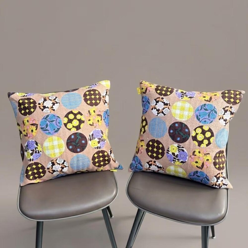K&ouml;nnte beinhalten: Zwei quadratische Kissen mit einem beigen Grundstoff, der mit einem sich wiederholenden Muster aus bunten Kreisen verziert ist. Die Kreise enthalten verschiedene Designs, darunter gelbe Blumen, blaue Streifen, braune Gitter und violette Blumenmotive.