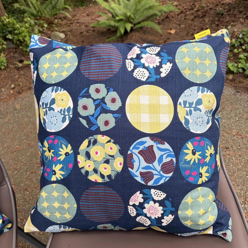 May include: A square throw pillow featuring a dark blue background with a repeating pattern of circles. Each circle contains different designs, including yellow and blue floral prints, yellow gingham checks, and dark blue horizontal stripes.