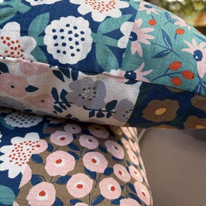K&ouml;nnte beinhalten: Ein Dekokissen mit einem Patchwork-Design aus Blumenmustern in Marineblau, Hellrosa, Salbeigr&uuml;n und Braun. Der Stoff zeigt verschiedene stilisierte Blumenformen, darunter runde Bl&uuml;ten und kleine Beeren, mit einer deutlich sichtbaren Webstruktur.