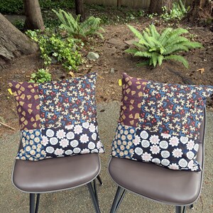 May include: Two square throw pillows featuring a patchwork design of dark fabric panels. The patterns include a dark purple section with yellow tulip-like shapes, a dark floral print with cream flowers, and a blue section with white circular motifs.