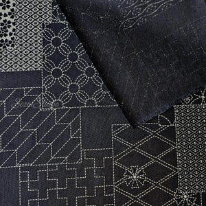 May include: A dark navy blue fabric featuring a patchwork design of white geometric patterns and floral motifs reminiscent of sashiko stitching. The textile displays various squares with intricate lines, crosses, and circles, with the text Sozai Studio.