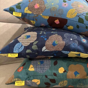 Puede incluir: Tres cojines cuadrados apilados verticalmente, cada uno con un patr&oacute;n floral de grandes flores estilizadas en tonos azul, marr&oacute;n, amarillo y verde. Cada coj&iacute;n tiene una peque&ntilde;a etiqueta amarilla que dice Atelier Dorothy Japanese Fabric.