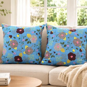 Puede incluir: Dos cojines cuadrados con un fondo azul brillante y un patr&oacute;n floral repetitivo. El dise&ntilde;o incluye flores estilizadas en tonos marr&oacute;n oscuro, gris y amarillo, acentuadas por peque&ntilde;as hojas verdes y tallos delgados sobre una tela azul claro.