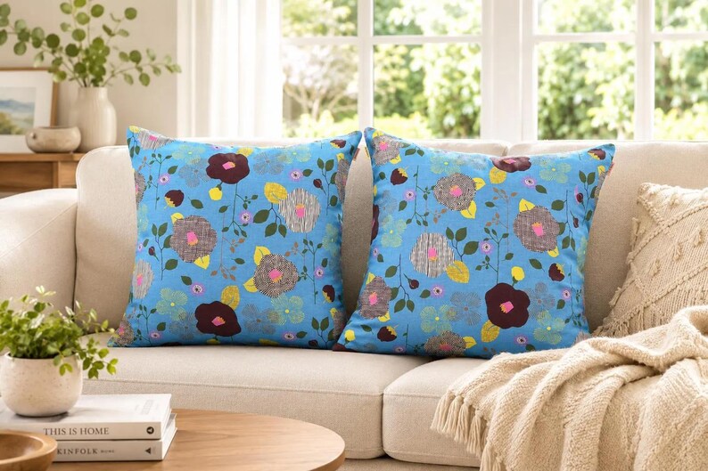 Puede incluir: Dos cojines cuadrados con un fondo azul brillante y un patr&oacute;n floral repetitivo. El dise&ntilde;o incluye flores estilizadas en tonos marr&oacute;n oscuro, gris y amarillo, acentuadas por peque&ntilde;as hojas verdes y tallos delgados sobre una tela azul claro.