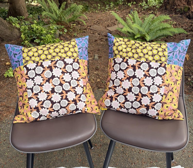 Peut inclure: Deux coussins carr&eacute;s pr&eacute;sentant un motif patchwork de tissus &agrave; fleurs. Les motifs incluent des fleurs jaunes sur fond sombre, des imprim&eacute;s floraux violet clair et un panneau central de fleurs blanches et roses sur fond brun fonc&eacute;.