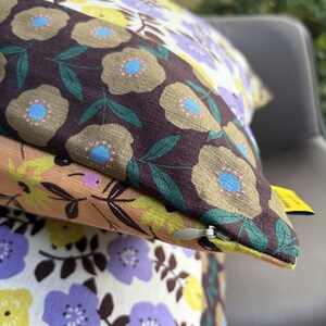 Peut inclure: Un coussin carr&eacute; orn&eacute; d'un motif floral avec des fleurs marron fonc&eacute;, beige, violet clair et jaune sur un tissu textur&eacute;. Le coussin pr&eacute;sente une fermeture &eacute;clair dissimul&eacute;e avec une tirette argent&eacute;e et une petite &eacute;tiquette jaune sur le c&ocirc;t&eacute;.