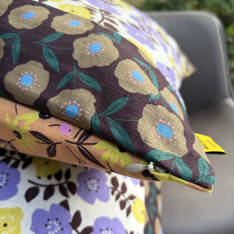Peut inclure: Un coussin carr&eacute; orn&eacute; d'un motif floral avec des fleurs marron fonc&eacute;, beige, violet clair et jaune sur un tissu textur&eacute;. Le coussin pr&eacute;sente une fermeture &eacute;clair dissimul&eacute;e avec une tirette argent&eacute;e et une petite &eacute;tiquette jaune sur le c&ocirc;t&eacute;.