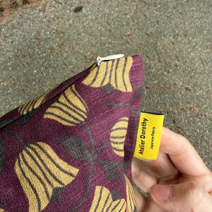 May include: A purple fabric cushion cover featuring a repeating pattern of golden yellow tulip-like flowers and dark gray stems. A white zipper is visible at the top edge. A small yellow tag is attached to the side with the text Atelier Dorothy Japanese Fabric.