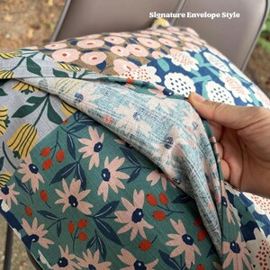 K&ouml;nnte beinhalten: Ein dekorativer Kissenbezug mit einem Verschluss im Umschlagstil. Der Stoff zeigt ein mehrfarbiges Blumenmuster mit rosa, gelben und wei&szlig;en Bl&uuml;ten auf blaugr&uuml;nem, blauem und grauem Hintergrund, akzentuiert durch kleine orangefarbene Beeren.