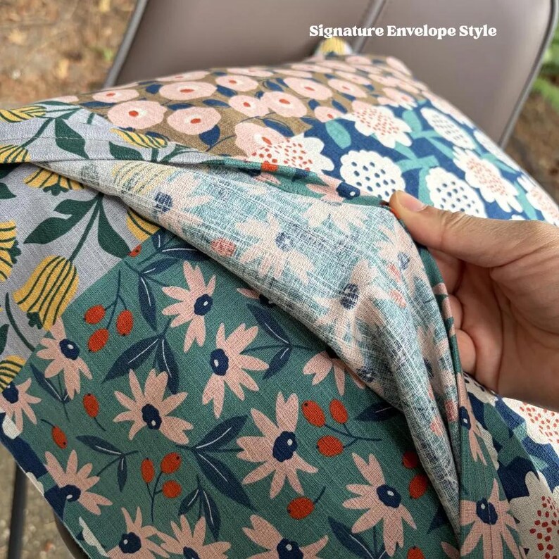 K&ouml;nnte beinhalten: Ein dekorativer Kissenbezug mit einem Verschluss im Umschlagstil. Der Stoff zeigt ein mehrfarbiges Blumenmuster mit rosa, gelben und wei&szlig;en Bl&uuml;ten auf blaugr&uuml;nem, blauem und grauem Hintergrund, akzentuiert durch kleine orangefarbene Beeren.