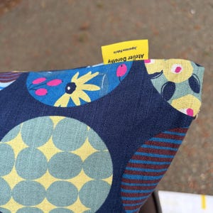 May include: A fabric cushion cover featuring a dark blue base with a pattern of large circles containing yellow, light blue, and pink floral and geometric designs. A small yellow tag is attached to the edge with the text Atelier Dorothy Japanese Fabric.