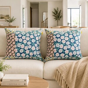 K&ouml;nnte beinhalten: Zwei quadratische Dekokissen mit einem Patchwork-Design, bestehend aus einem dunkelblauen Blumenmuster auf dem Hauptteil und einem kleineren braun-wei&szlig;en Blumenmuster auf dem Seitenteil, jeweils mit einem kleinen gelben Etikett am rechten Rand.