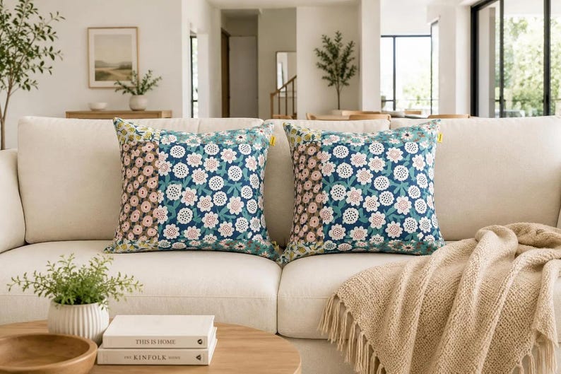K&ouml;nnte beinhalten: Zwei quadratische Dekokissen mit einem Patchwork-Design, bestehend aus einem dunkelblauen Blumenmuster auf dem Hauptteil und einem kleineren braun-wei&szlig;en Blumenmuster auf dem Seitenteil, jeweils mit einem kleinen gelben Etikett am rechten Rand.
