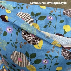Puede incluir: Una funda de coj&iacute;n azul claro con un estampado floral de motivos circulares amarillos, morados y rayados en blanco y negro, hojas verdes y tallos marrones finos. La tela tiene un tejido visible y el texto Signature Envelope Style.