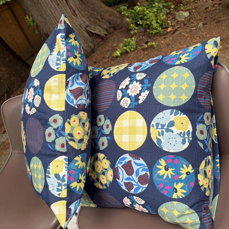 May include: Two square throw pillows with a dark navy blue background featuring a repeating pattern of colorful circles. Each circle contains distinct designs including yellow gingham, floral arrangements, and striped textures in blue, yellow, and green.