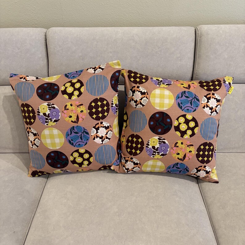 K&ouml;nnte beinhalten: Zwei quadratische Kissen mit einem hellbraunen Grundstoff, der mit einem sich wiederholenden Muster aus bunten Kreisen verziert ist. Jeder Kreis enth&auml;lt verschiedene Designs, darunter Blumenmuster, gelb-wei&szlig;es Vichy-Karo und dunkelbraune Punkte.